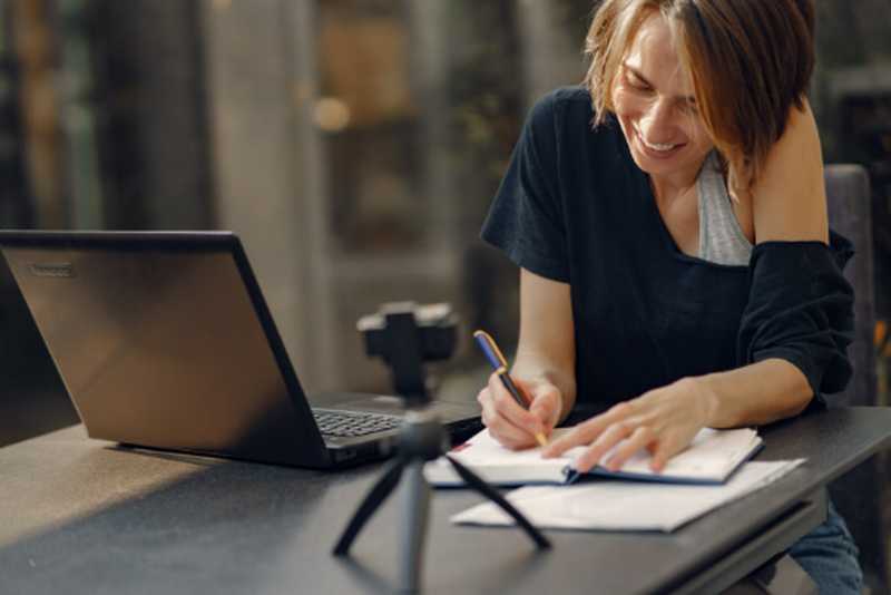 Eine Frau sitzt vorm Laptop und schreibt in ein Notizbuch